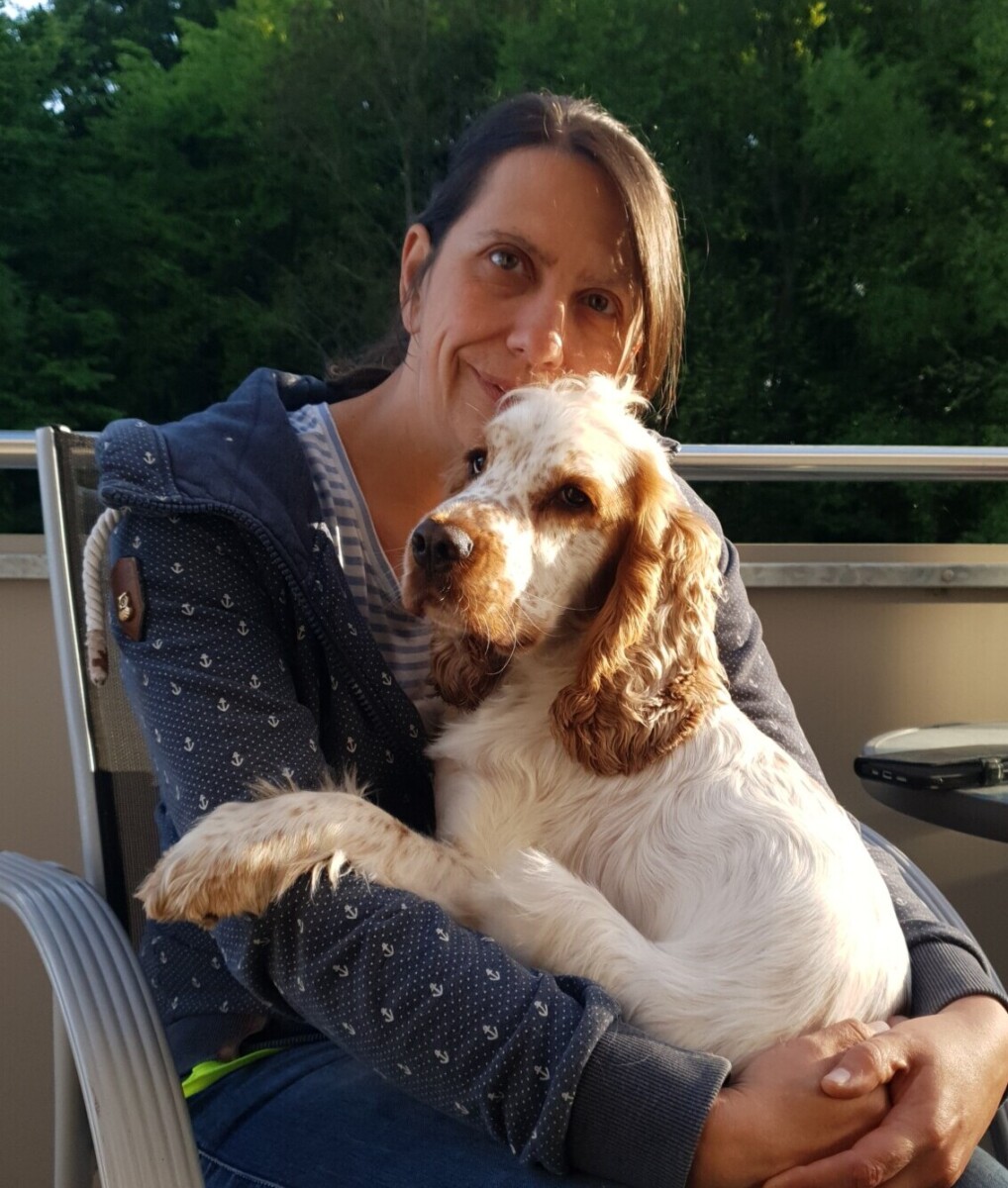 Gründerin von Ruhrpawt sitzt auf einem Stuhl auf einem Balkon mit einem roan orange Cocker Spaniel auf dem Schoß und lächelt in die Kamera